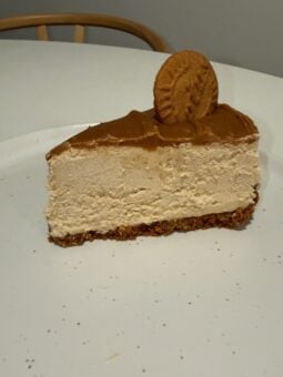 A slice of vegan Biscoff cheesecake topped with caramel and a round biscuit on a white plate.