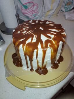 A round vegan pumpkin cake with white cream cheese frosting, topped with a generous drizzle of caramel sauce that drips down the sides onto a yellow cake stand.