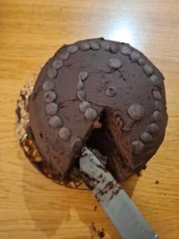 A round vegan gluten-free chocolate cake topped with chocolate drops, with a slice removed by a cake server.