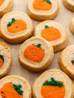 Round sugar cookies featuring a centered orange pumpkin design with a small green stem, arranged on parchment paper.