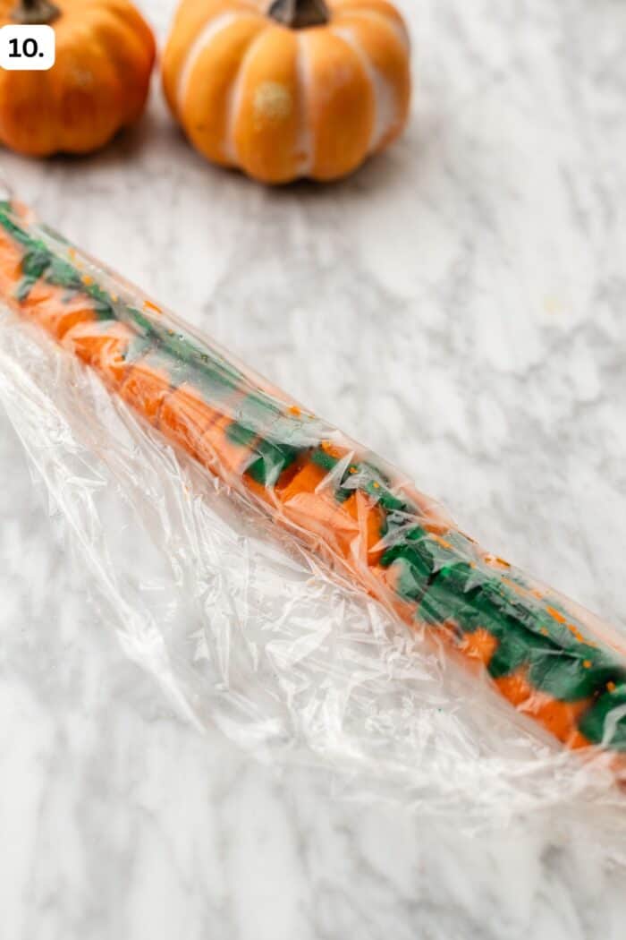 A log of orange and green cookie dough wrapped in plastic film, with two small pumpkins in the background.