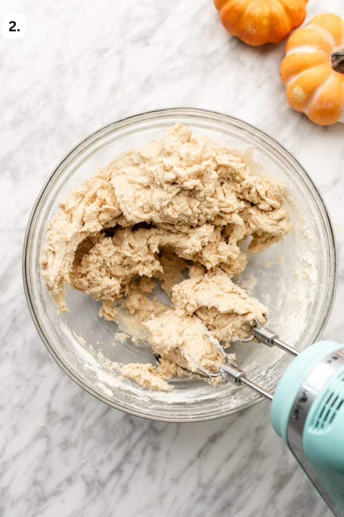 A glass bowl containing thick cookie dough being mixed by a handheld electric mixer, with two small pumpkins sitting on the marble surface nearby.