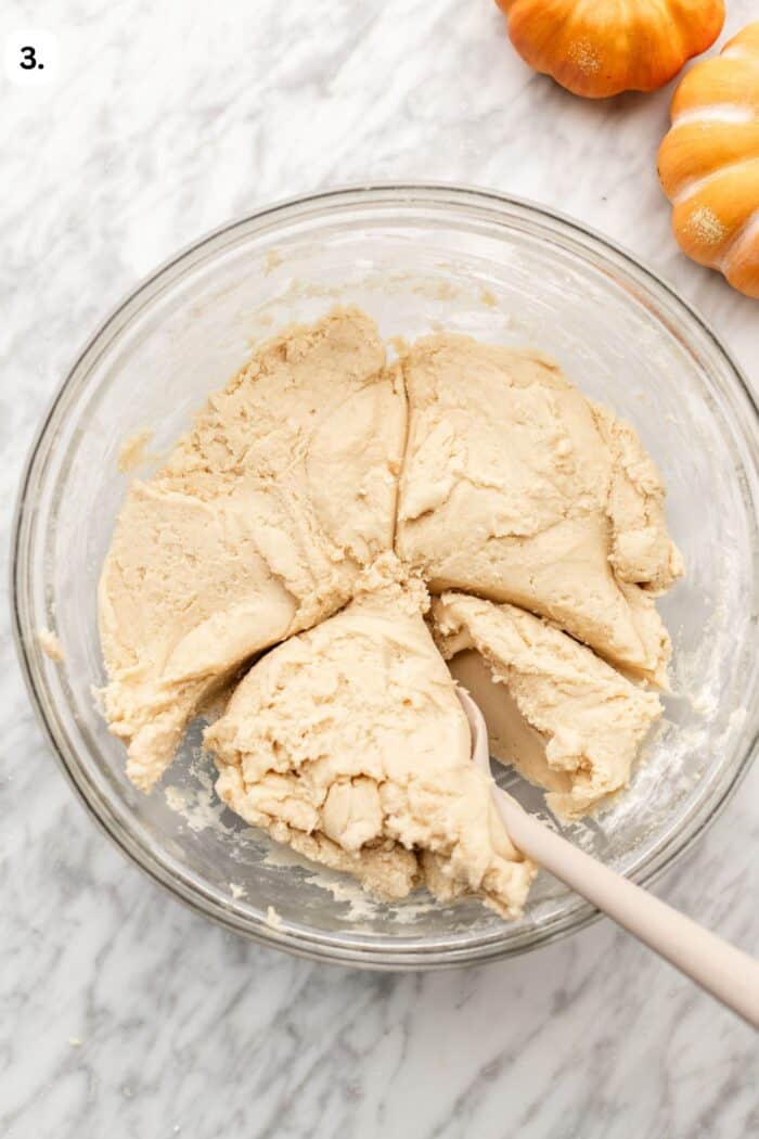 A glass bowl containing a large mound of pale dough being portioned with a white spatula, with two small orange pumpkins nearby.