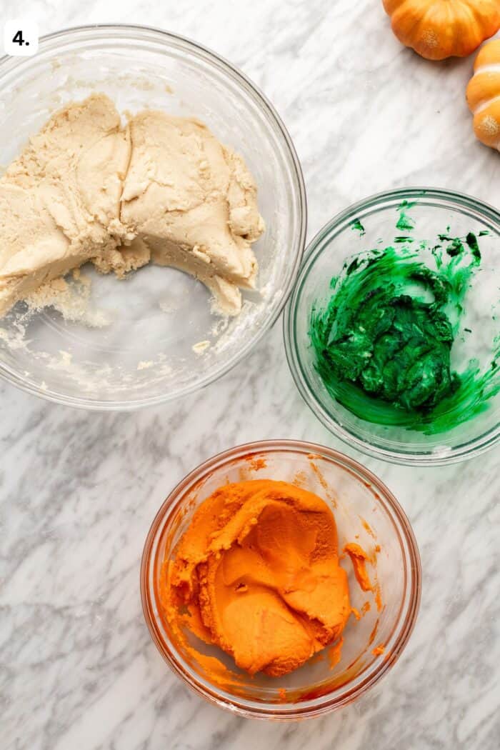 Three glass bowls on a marble surface: one containing plain beige dough, one with green-dyed dough, and one with orange-dyed dough.