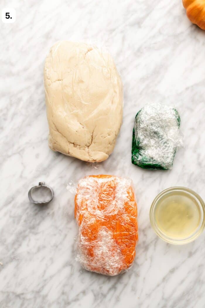 Three blocks of dough—large beige, small green, and medium orange—wrapped in plastic film, next to a pumpkin-shaped cookie cutter and a small bowl of liquid on a marble surface.