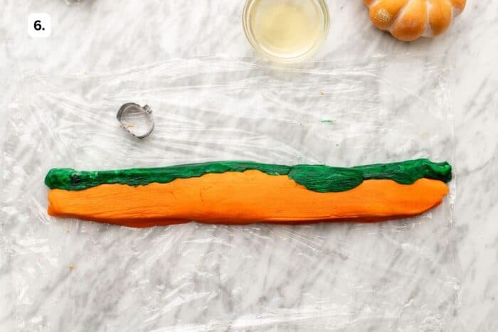 A log of orange and green dough on plastic wrap, next to a small pumpkin-shaped cookie cutter and a bowl of liquid.