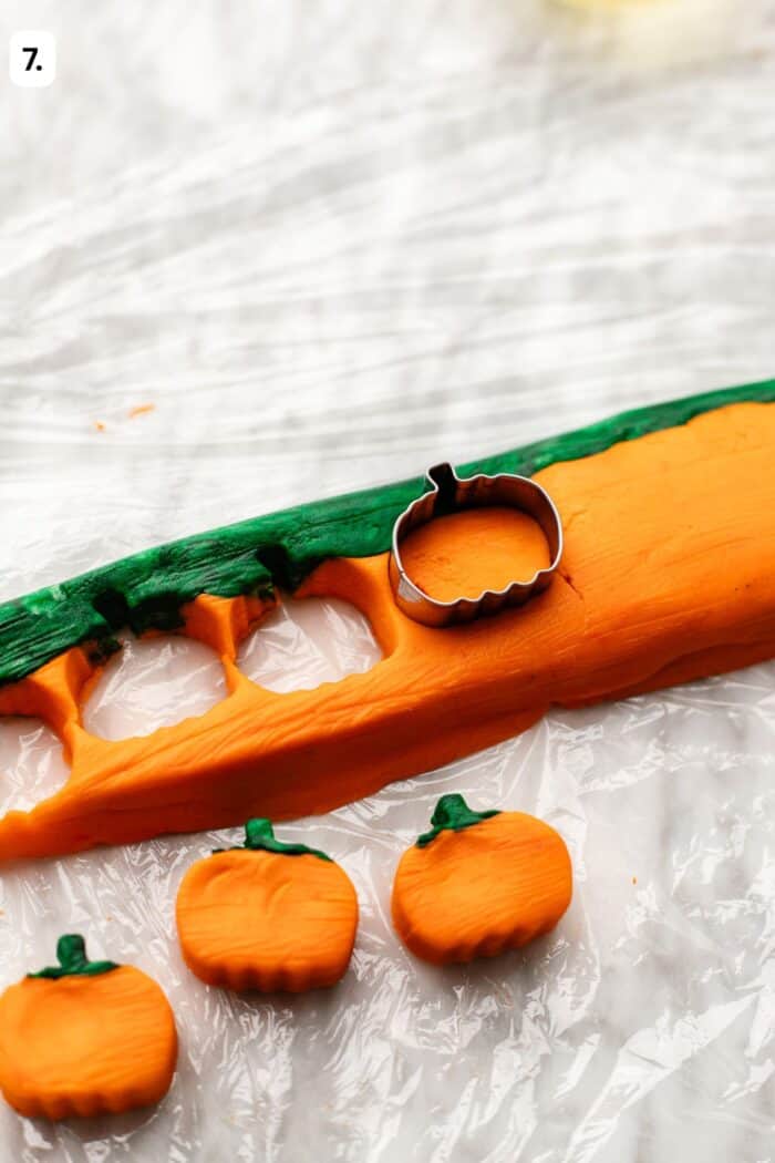 A metal pumpkin-shaped cookie cutter sits on a flat piece of orange and green dough, with three small pumpkin-shaped cutouts below.