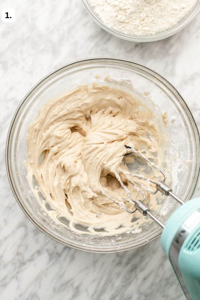 A handheld electric mixer blending batter in a glass bowl, with a small bowl of flour nearby.