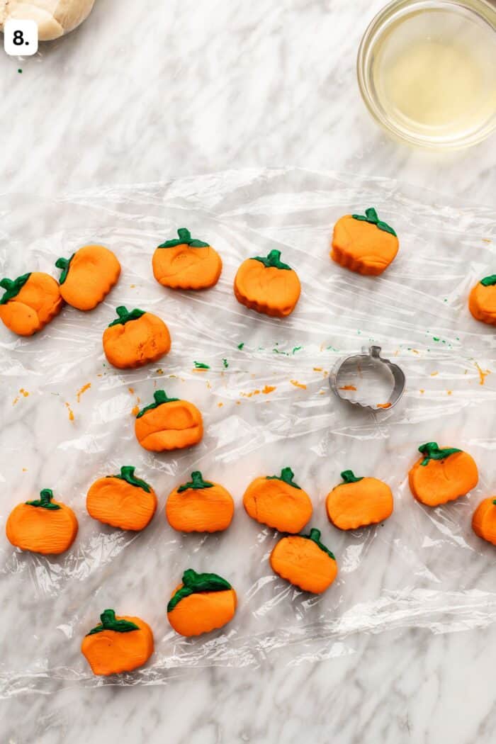 Small orange pumpkin-shaped cookies with green stems scattered on plastic wrap, with a metal pumpkin cookie cutter nearby.