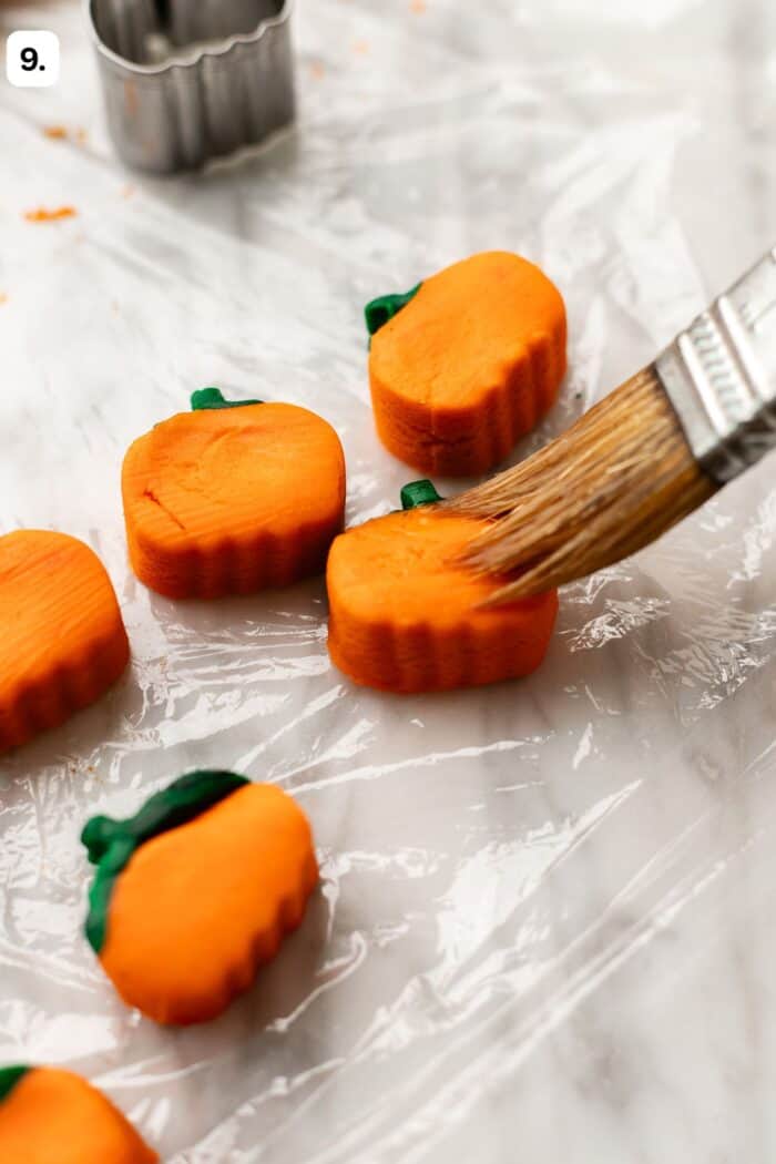 A pastry brush glazes small, orange pumpkin-shaped cookies on a sheet of plastic wrap.