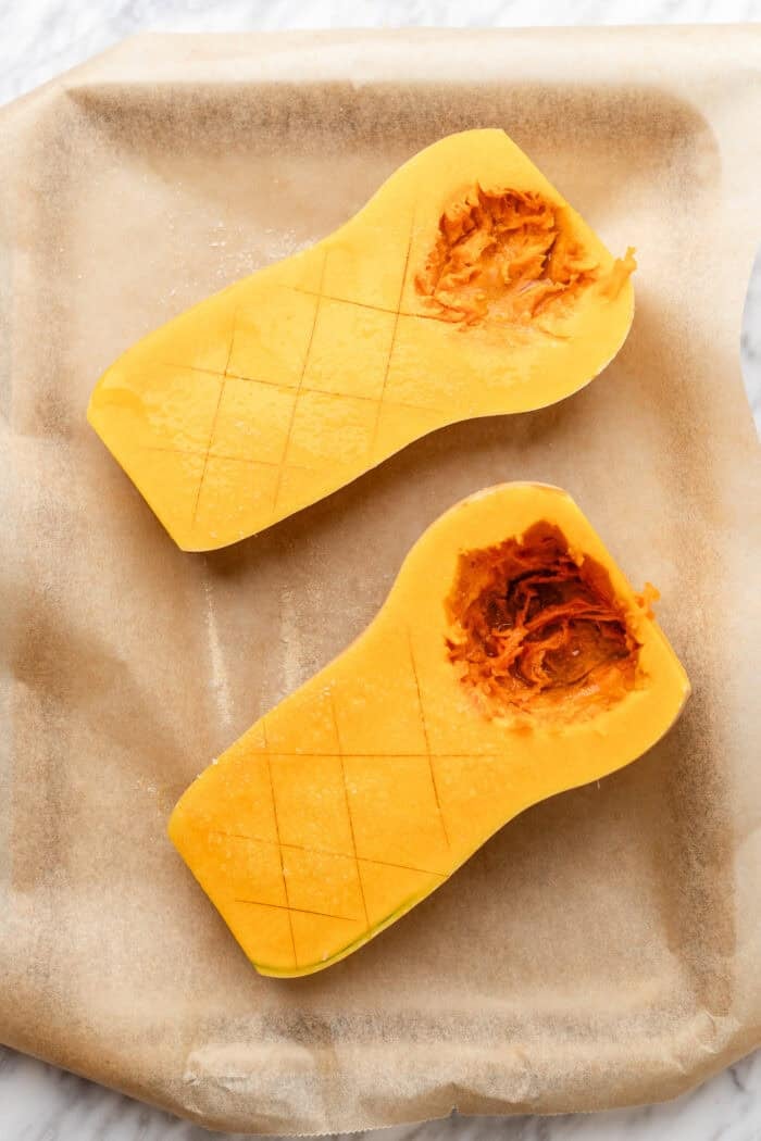Two halves of a butternut squash with seeds removed and flesh scored in a crisscross pattern, placed on parchment paper on a baking sheet.