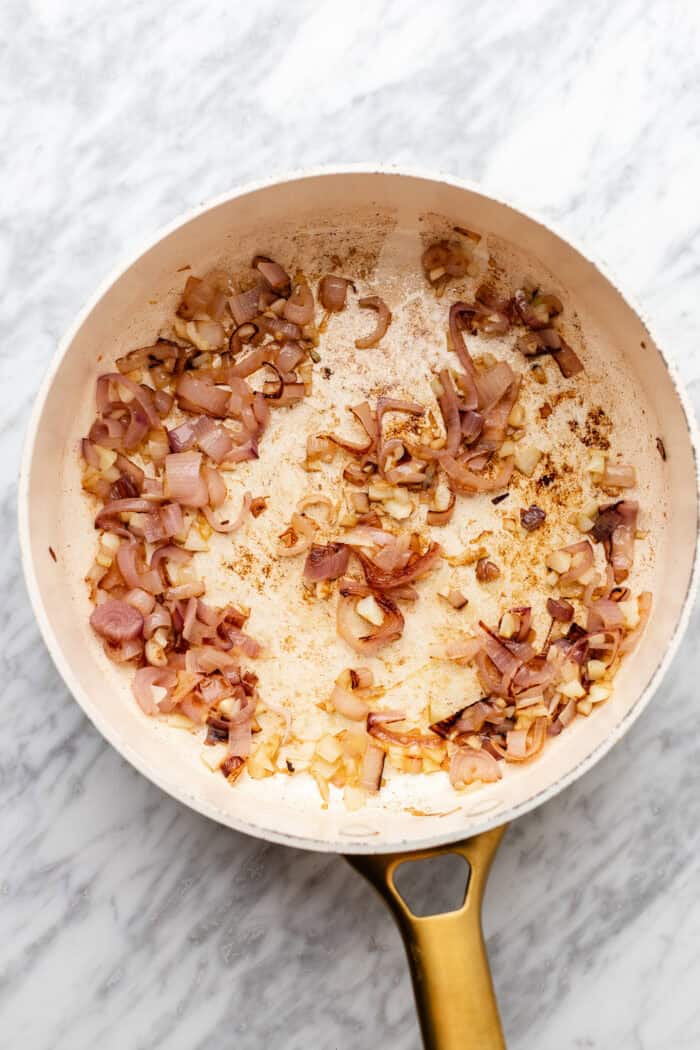 A white skillet with sautéed chopped onions and garlic, some pieces browned, sits on a light marble surface.