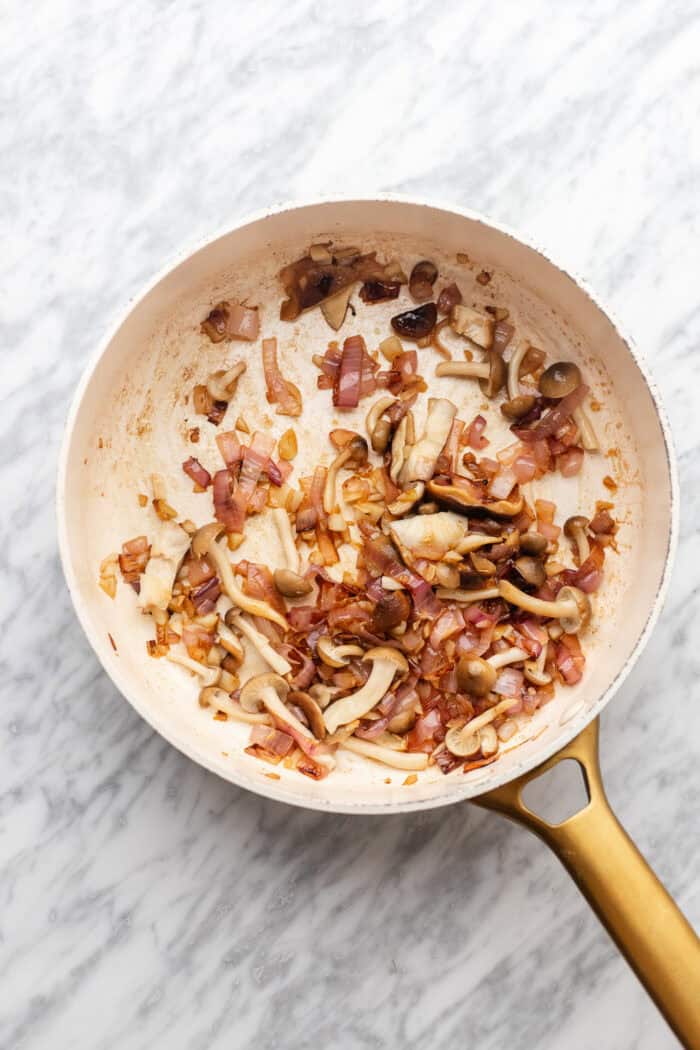 A white skillet with a gold handle contains sautéed mushrooms and diced red onions on a marble surface.