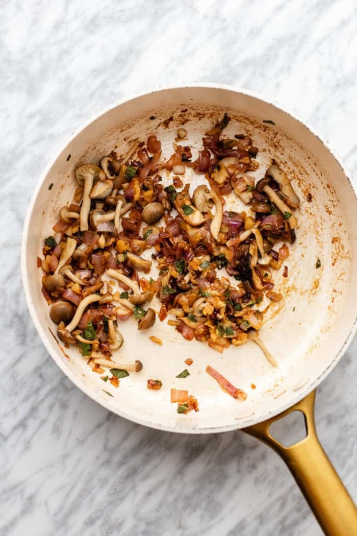 A white skillet with sautéed mushrooms, onions, and herbs sits on a marble surface. The vegetables are browned and mixed together, with a gold handle on the pan.