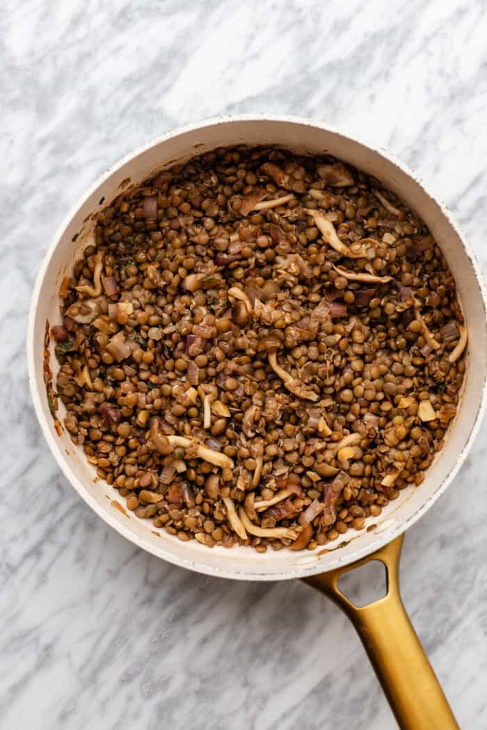 A white pan with a gold handle contains cooked lentils mixed with sliced mushrooms and diced onions, sitting on a marble countertop.