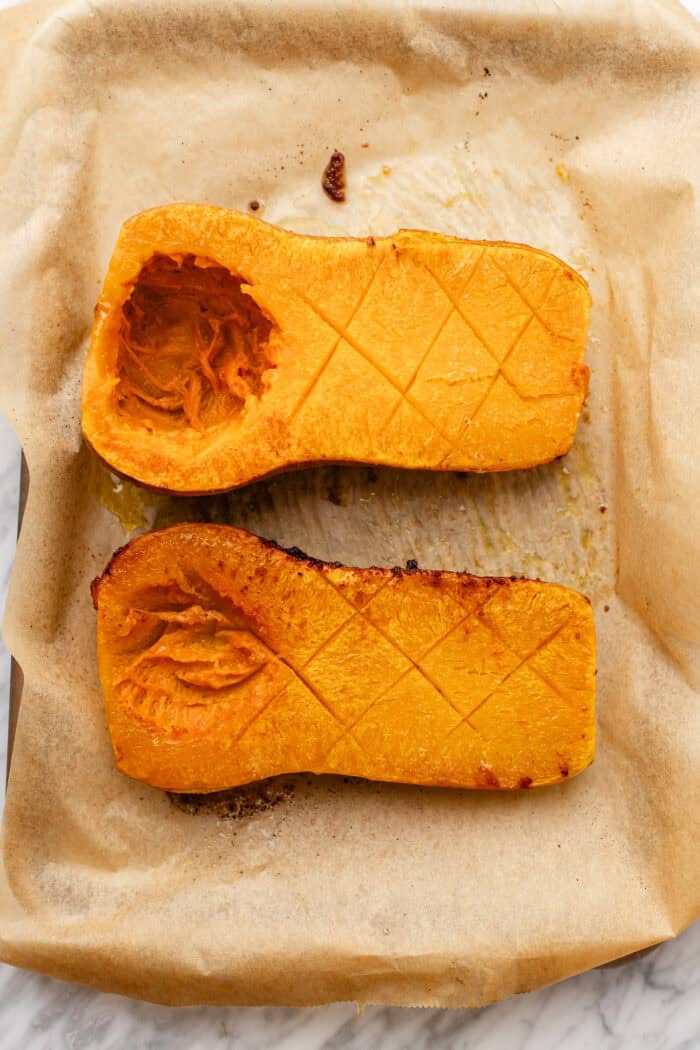 Two halves of roasted butternut squash with criss-cross scoring on the flesh, placed on a sheet of parchment paper atop a baking sheet.