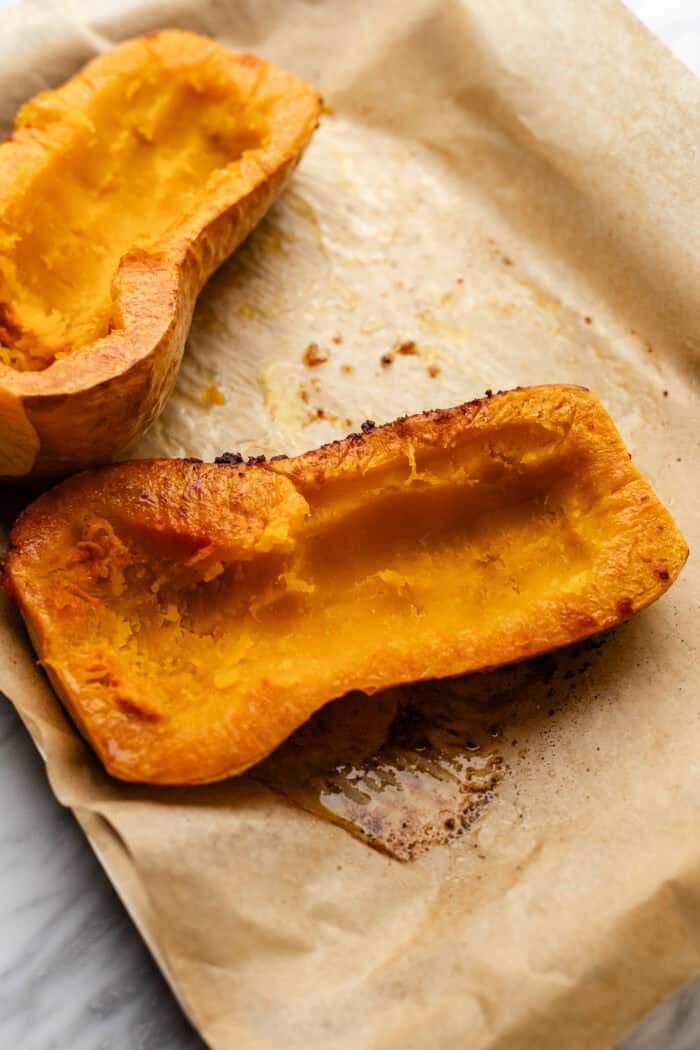 Two roasted butternut squash halves with golden, caramelized edges rest on a sheet of parchment paper, showing their soft, cooked interiors.