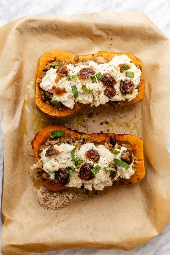 Two halves of roasted butternut squash topped with vegan cheese, herbs, and chopped dates, before baking on parchment paper on a baking tray.