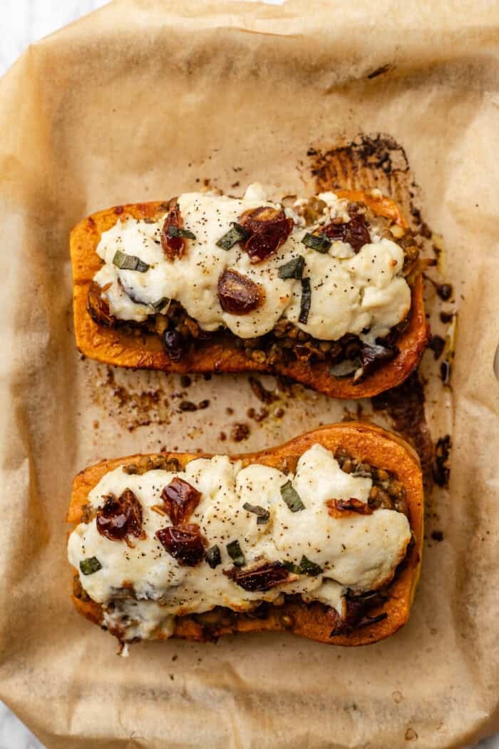 Two halves of roasted butternut squash filled with a savory mixture, topped with melted cheese, herbs, and chopped dates, on parchment paper.