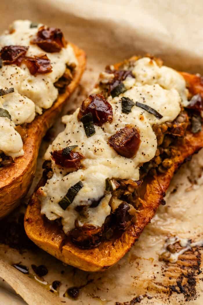 Two halves of roasted butternut squash stuffed with a savory filling, topped with melted cheese and herbs, resting on parchment paper.