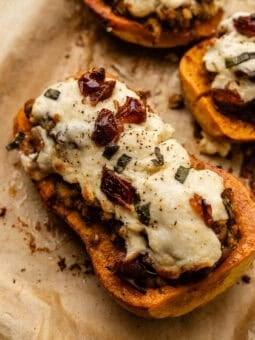 A roasted butternut squash half stuffed with a lentil mixture, topped with melted cheese, herbs, and bits of sun-dried tomato, resting on parchment paper.