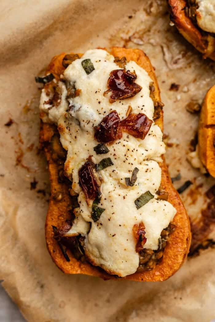 A roasted half of butternut squash stuffed with vegetables, topped with melted cheese and herbs, on parchment paper.