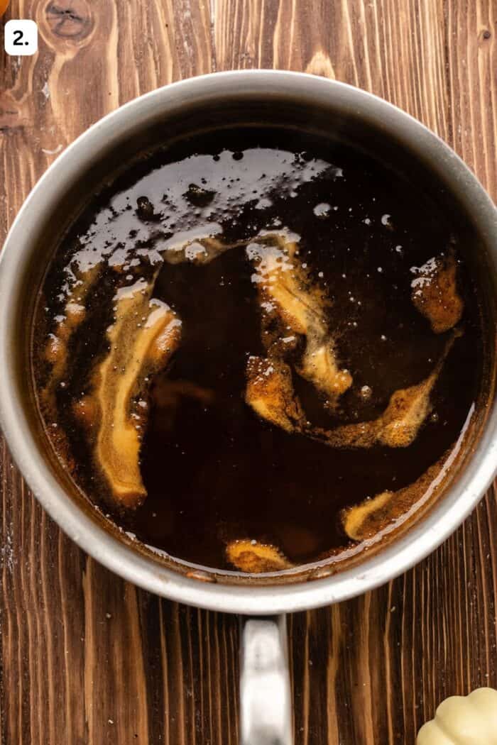 A metal saucepan filled with dark, simmering liquid with swirls of golden fat on the surface.