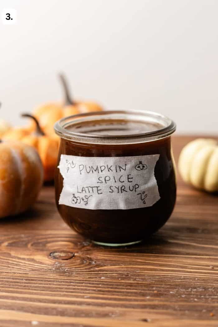 A glass jar of dark brown syrup labeled Pumpkin Spice Latte Syrup with hand-drawn pumpkins, set on a wooden table with small pumpkins in the background.