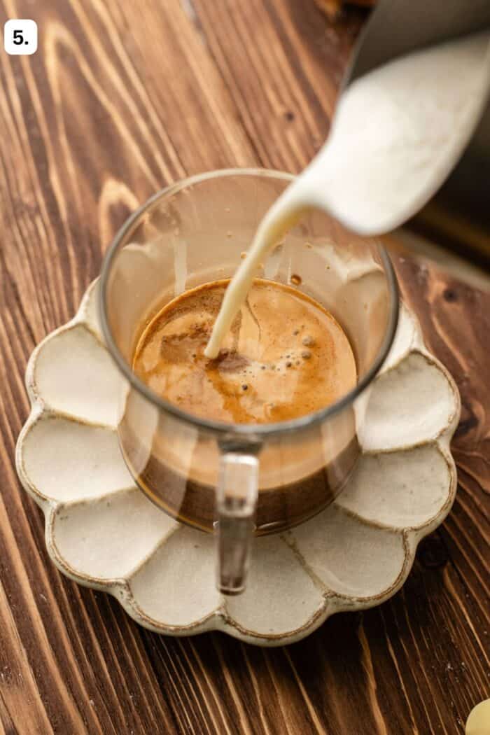 Milk being poured from a pitcher into a glass cup of espresso sitting on a scalloped saucer.