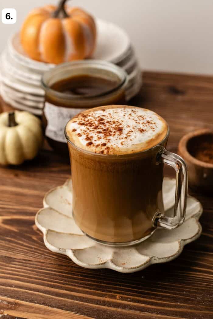 A glass mug of latte topped with foam and cinnamon on a scalloped saucer, with small decorative pumpkins in the background.