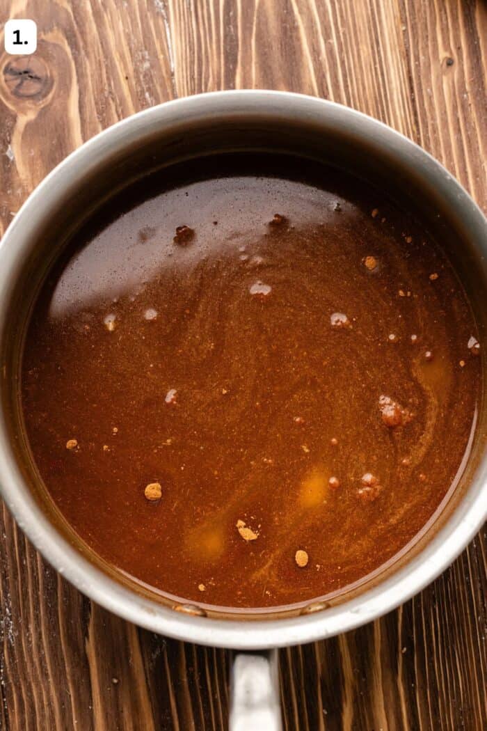 A saucepan containing a dark brown liquid sauce with visible spices on a wooden surface.