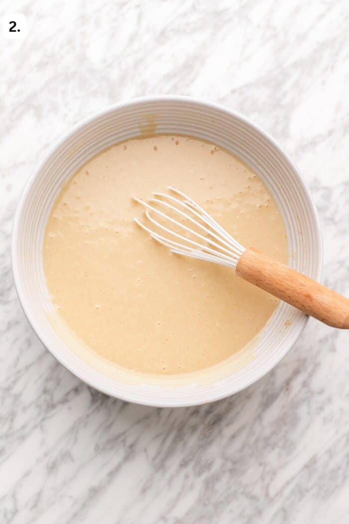 A wire whisk rests in a bowl of smooth, light brown batter on a marble countertop.