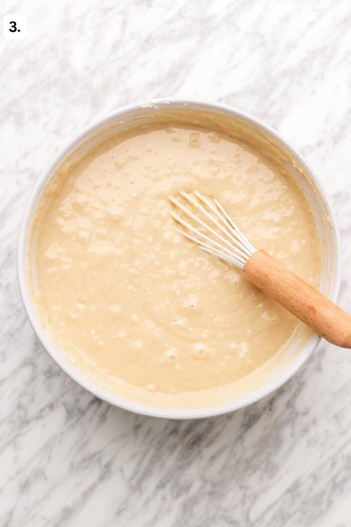 A whisk rests in a bowl of smooth, light-tan cake batter.