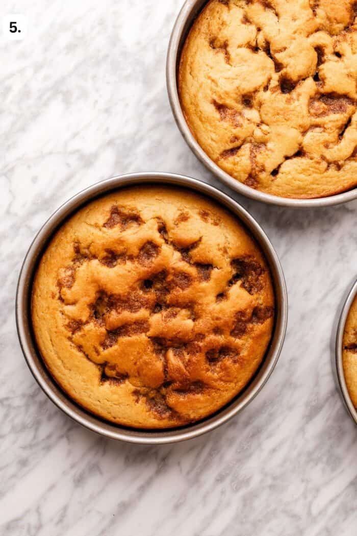Two round cinnamon swirl cakes in metal baking pans on a marble surface.