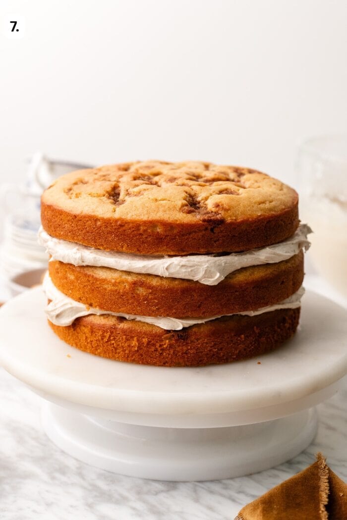 A three-layer cinnamon cake with frosting between each layer on a marble cake stand.