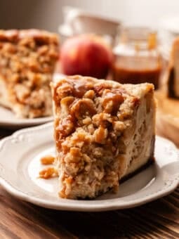 slice of vegan apple crumble cheesecake on a white plate with an apple and caramel jar in the background all on a wooden table.