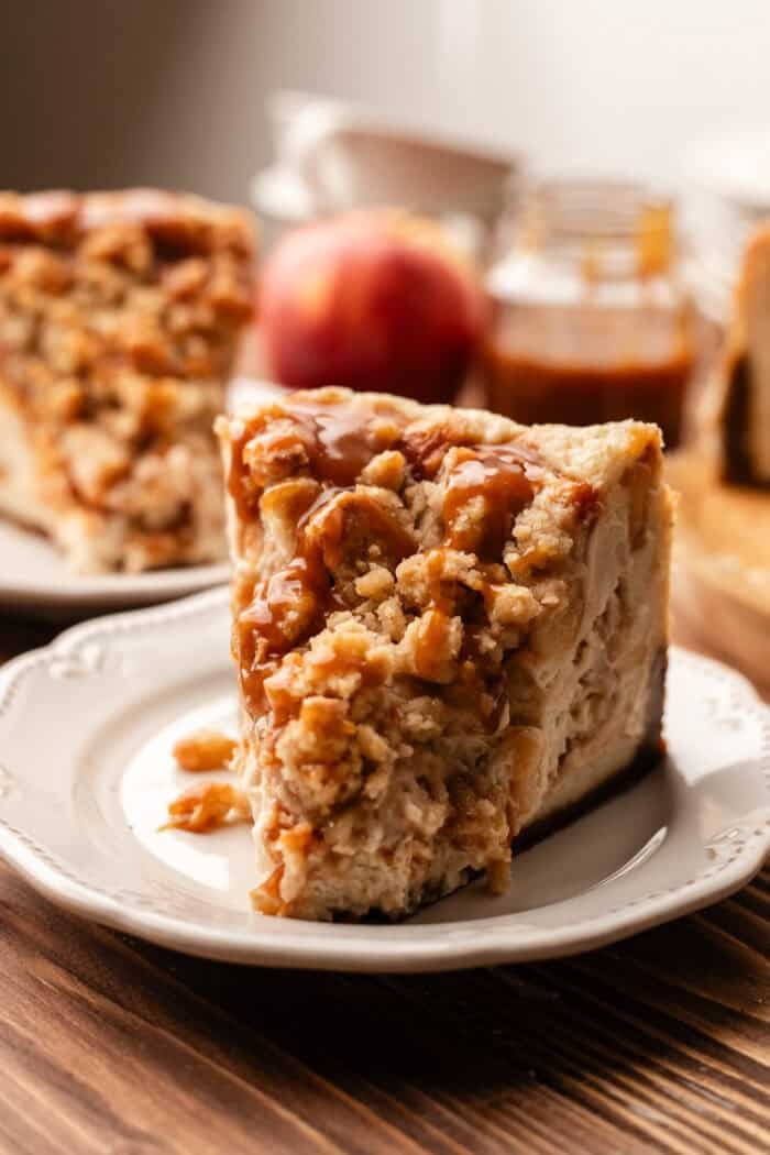 slice of vegan apple crumble cheesecake on a white plate with an apple and caramel jar in the background all on a wooden table.