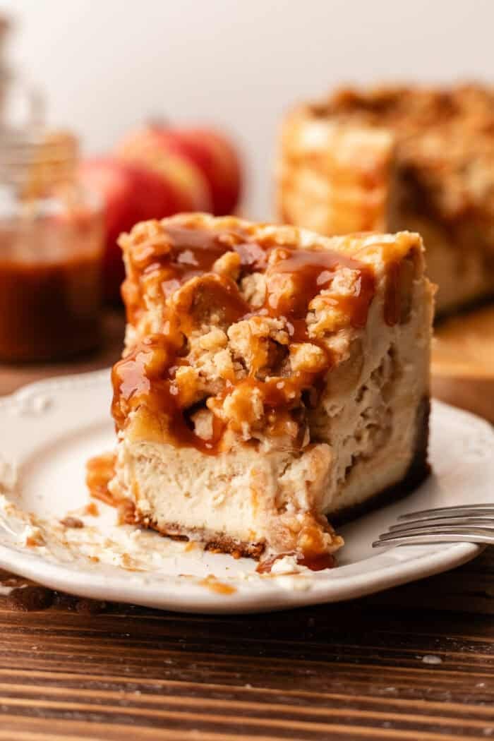 bitten slice of apple crumble cheesecake on a white plate with apples in the background as well as the sliced cheesecake and caramel jar on a wooden table.