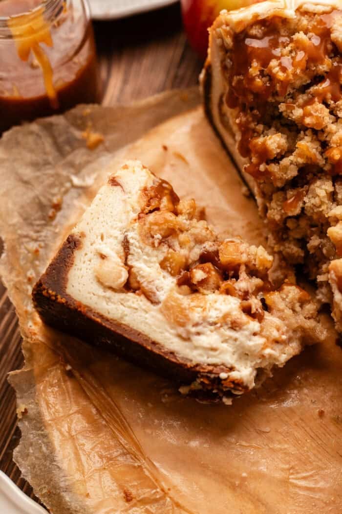 sliced vegan apple crumble cheesecake on the cutting board with the full cheesecake next to it and a jar of caramel sauce.