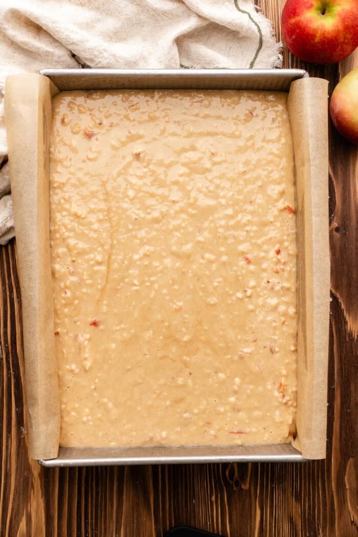 apple cake batter poured into a 9x13" pan on a wooden table with an apple next to it.