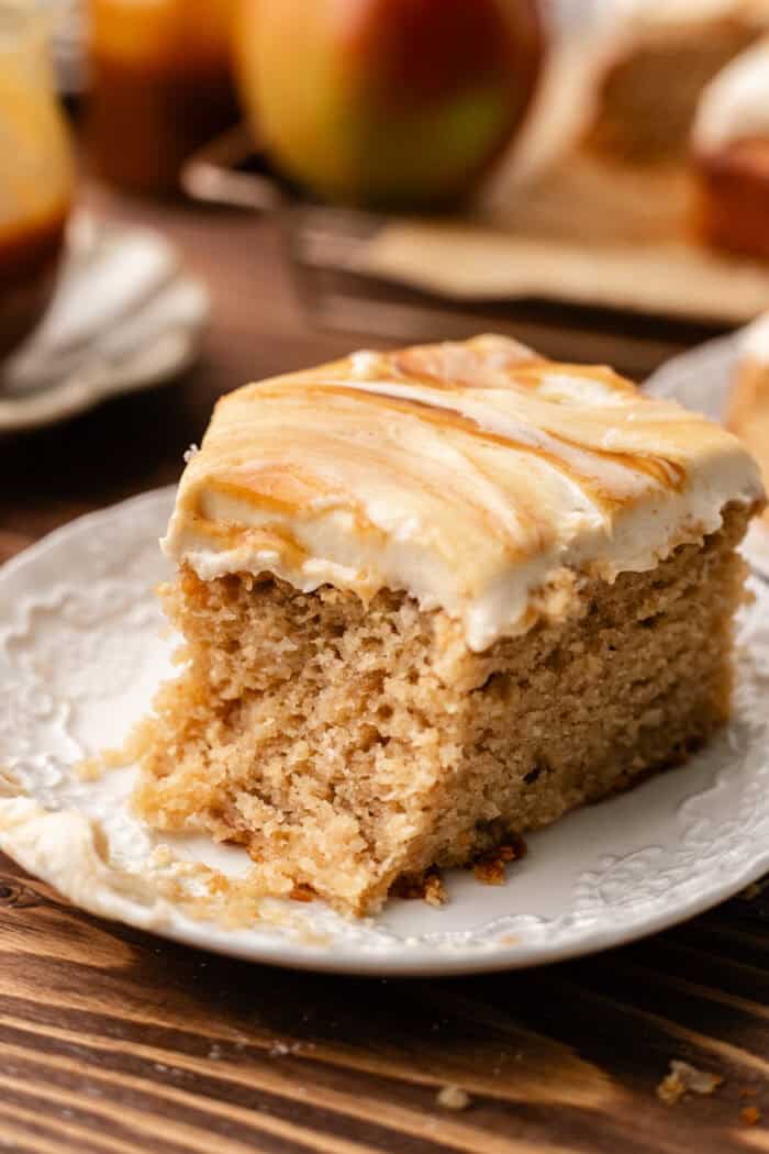 bitten slice of apple spice cake on a white plate with apples and caramel jars behind it.