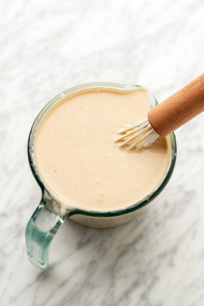 A glass measuring cup filled with creamy beige sauce being whisked with a wooden-handled whisk, on a white marble surface.