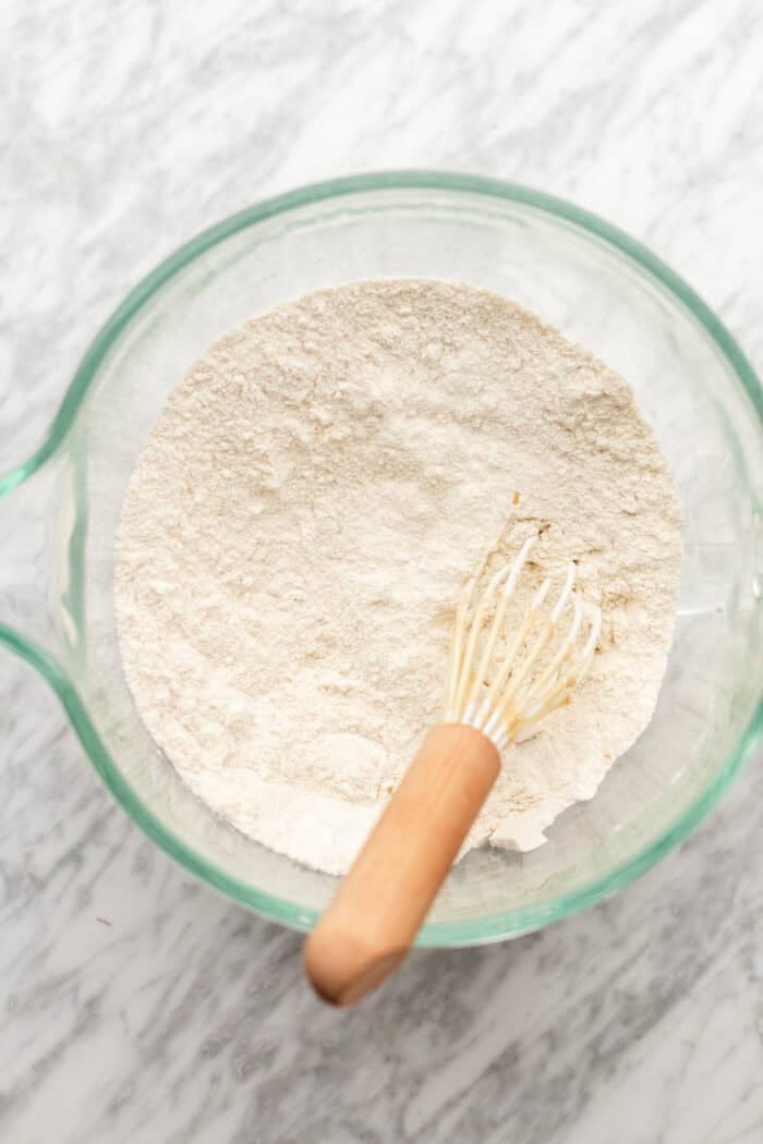 A glass bowl filled with flour and a small whisk with a wooden handle, placed on a marble countertop.