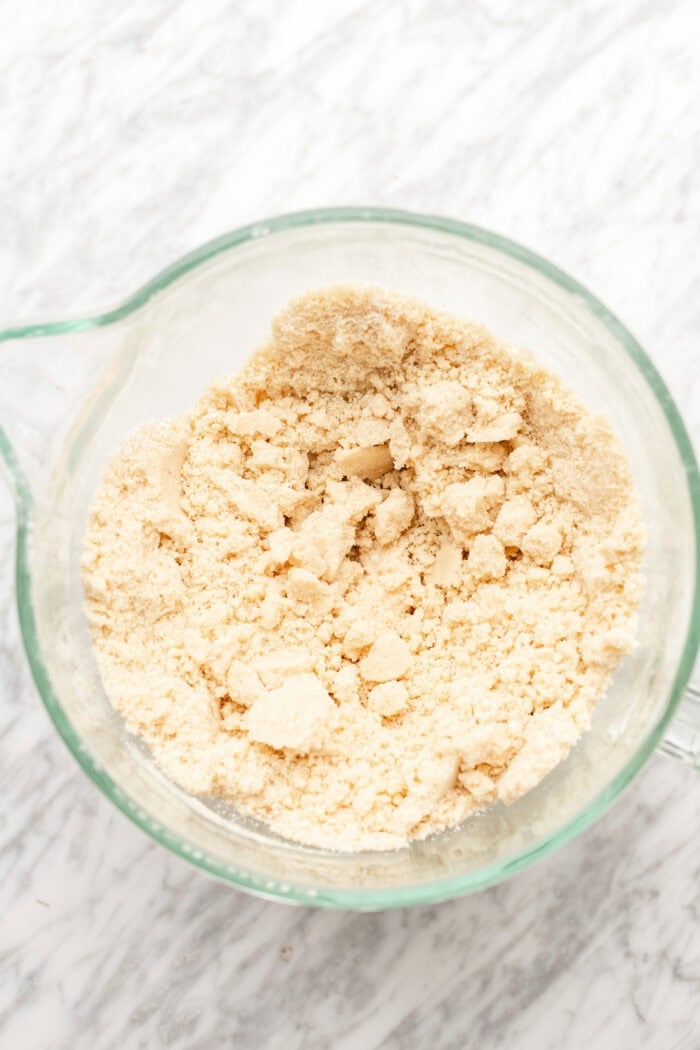A glass mixing bowl filled with coarse, crumbly dough mixture sits on a white marble surface.