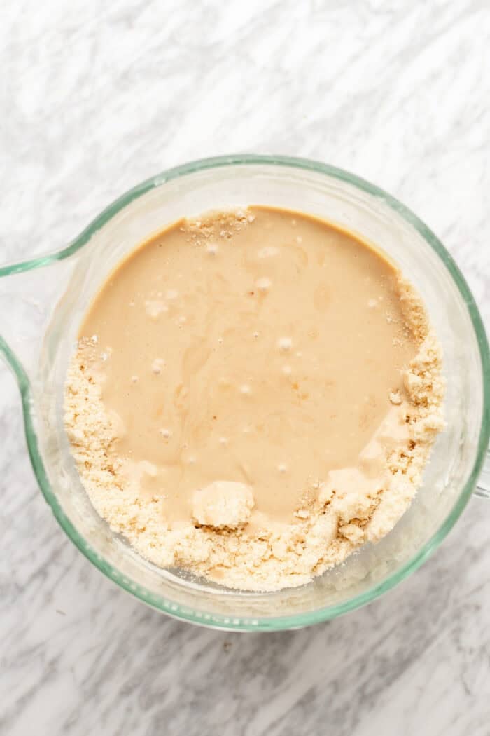 A glass mixing bowl with flour and a tan liquid partially mixed together, sitting on a white marble surface.