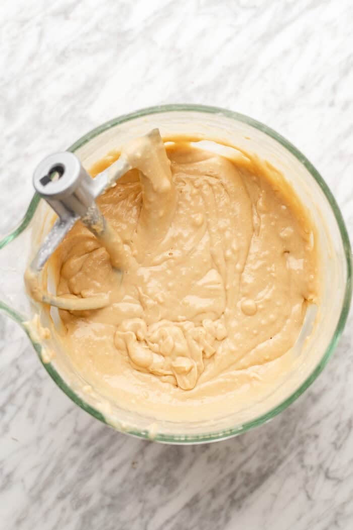 A glass mixing bowl filled with light tan cake batter sits on a marble countertop, with a stand mixer paddle attachment resting inside.