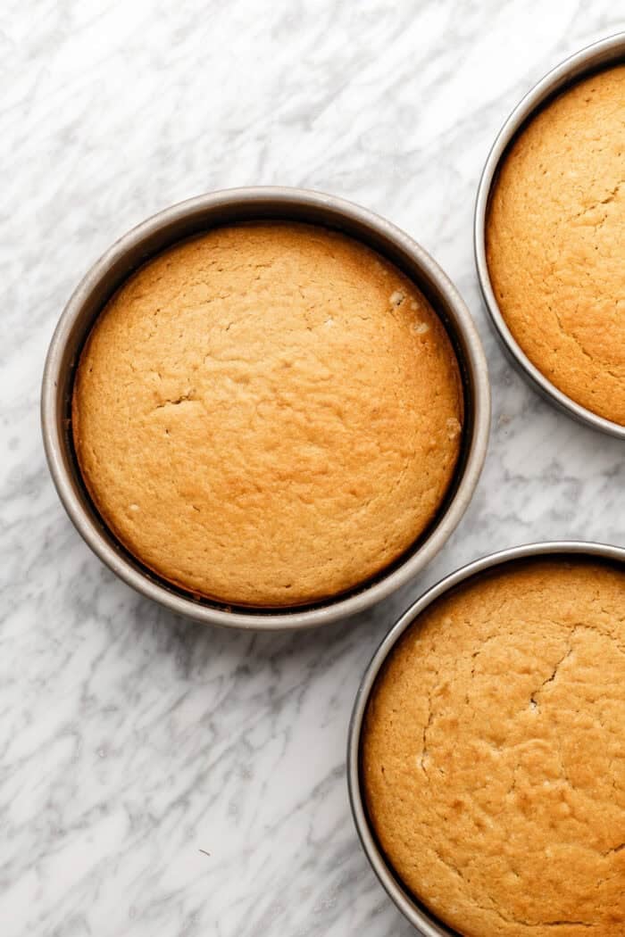 Three round, golden-brown cakes in metal pans are sitting on a white marble surface. The cakes have a smooth, even top and are arranged close together, with some overlapping in the image.