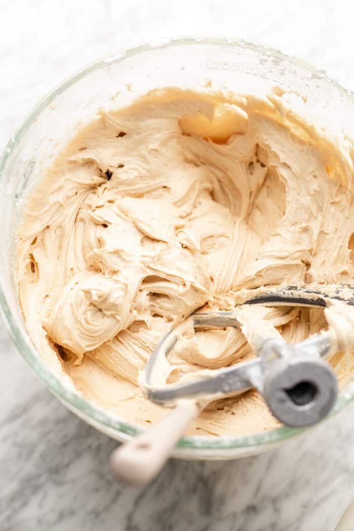 A glass mixing bowl filled with creamy, light tan cookie dough. A metal beater and spatula rest inside the bowl, which sits on a white marble surface.