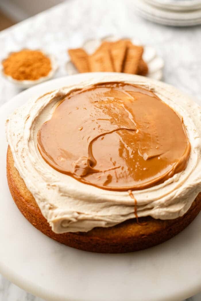 A round cake with a thick layer of creamy frosting and smooth caramel sauce on top, displayed on a white surface with cookies and brown sugar in the background.
