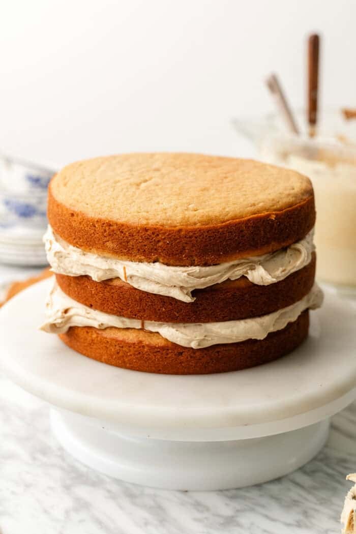 A three-layer round cake with light brown frosting between each layer sits on a white cake stand. The top is unfrosted, and baking tools are visible in the background.